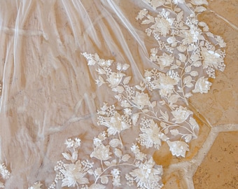 May include: A delicate, white bridal veil with intricate floral embroidery along the edge. The veil is made of a sheer fabric, allowing light to pass through. The floral design includes detailed flowers and leaves.