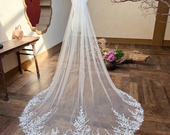 May include: A long, white bridal veil with intricate floral lace detailing along the bottom edge. The veil is displayed on a mannequin, showcasing its length and delicate design. The lace features a detailed floral pattern.