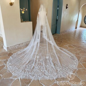 May include: A long, white, sheer tulle wedding veil featuring intricate, dense floral embroidery along the scalloped edges and a wide, ornate lace border that pools on the floor. The veil is draped over a stand, showcasing its length and detail.