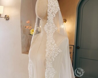 May include: A white bridal veil draped over a mannequin, featuring intricate floral lace embroidery along the edges. The sheer fabric flows downward, highlighting the detailed scalloped lace trim that frames the face and extends along the length.