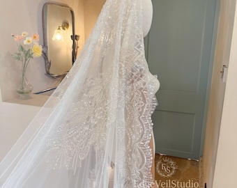 May include: A long white bridal veil featuring intricate silver embroidery and clear beadwork along the scalloped edges. The sheer fabric drapes over a mannequin, showcasing detailed floral patterns and sparkling embellishments throughout the design.