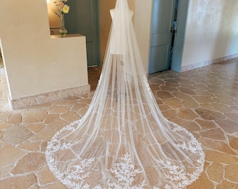 May include: A long, floor-length white bridal veil made of sheer tulle fabric. The veil features intricate white floral lace embroidery along the rounded hemline, creating a decorative border that spreads across the floor in a circular shape.