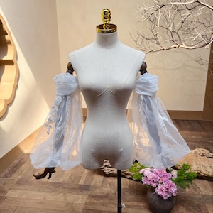 May include: A light-colored dress form displays a pair of sheer, white, off-the-shoulder sleeves. The sleeves are made of a delicate, flowing fabric with intricate lace detailing. The dress form has a gold-colored top and is set against a neutral background.