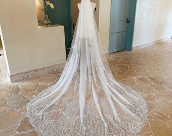 May include: A long, floor-length white bridal veil draped over a mannequin. The veil features sheer, lightweight tulle fabric with an intricate, dense floral lace embroidery pattern along the rounded hemline. The text EdgeVeilStudio is visible.