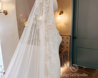 May include: A long, white bridal veil draped over a mannequin, featuring intricate floral lace trim along the edges. The sheer, lightweight fabric extends downward, showcasing a detailed lace pattern that frames the silhouette of the headpiece.