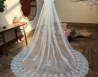 May include: A long, sheer white bridal veil with delicate blue floral embroidery along the edges. The veil is displayed on a mannequin, showcasing its length and intricate design. The floral details add a touch of elegance.