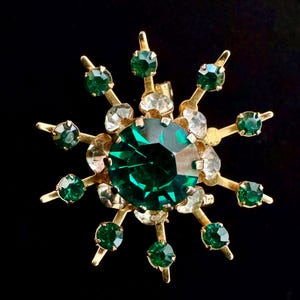 May include: A gold-toned brooch in a starburst design, featuring a large, faceted emerald-green center stone surrounded by smaller green and clear crystals. The brooch has a vintage aesthetic.