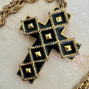 May include: A gold-tone necklace featuring a large cross pendant. The cross is black with gold diamond-shaped accents and gold studs. The chain is a thick, textured gold color.