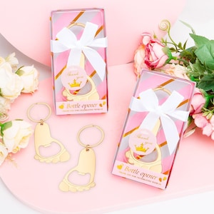 May include: Gold-colored bottle opener in the shape of a foot, with a keyring attached. The bottle opener is packaged in a clear box with a pink and gold striped design, tied with a white ribbon. The box has the text "Bottle opener" and "Thank you for celebrating with us."