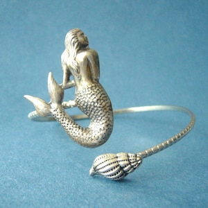 Peut inclure: Bracelet en argent avec une sirène et un charme en forme de coquillage. La sirène est représentée avec de longs cheveux flottants et une queue.