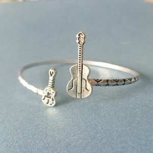 Könnte beinhalten: Silberfarbenes Armreif-Armband mit zwei Gitarren-Anhängern. Das Armband hat ein strukturiertes Design am Band und ist vor einem hellblauen Hintergrund platziert. Die Gitarren-Anhänger sind detailliert und fügen ein musikalisches Thema hinzu.