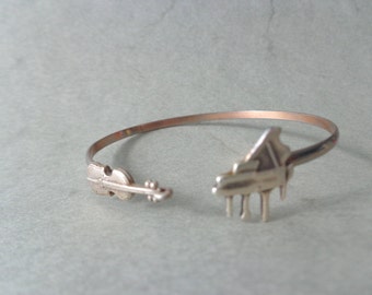 May include: A silver bracelet with a piano and violin design. The bracelet is open and has a simple, elegant design.