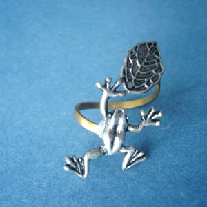 Puede incluir: Un anillo de plata con forma de rana con un diseño de hoja. La rana está sentada en una banda dorada y la hoja está en la espalda de la rana.
