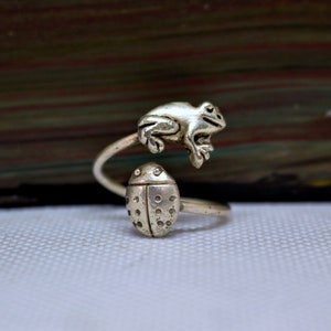 May include: A silver-toned ring featuring a frog and a ladybug design. The frog is perched above the finger, while the ladybug sits below. The ring has an adjustable, wrap-around style.