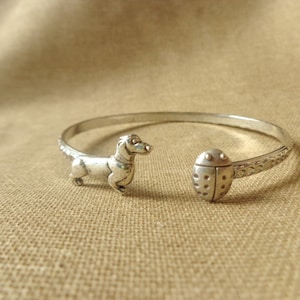 Puede incluir: Pulsera de plata con un encanto de dachshund y una mariquita. La pulsera está abierta y tiene un diseño texturizado.