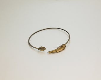 May include: An open cuff bracelet with a feather design. The bracelet is made of a thin, dark metal band. The feather design is gold-colored and detailed. The bracelet is open at one end.