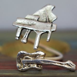 May include: A silver-toned ring featuring a detailed piano and guitar design. The ring is made of metal and has a unique, twisted shape.