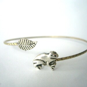 Könnte beinhalten: Ein silbernes Armreif-Armband mit einem silbernen Frosch-Anhänger und einem silbernen Blatt-Anhänger.