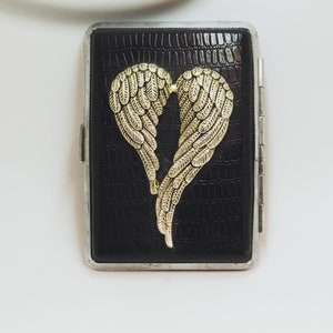 May include: Black cigarette case with a silver frame and a textured, faux-leather finish. The case features a gold-colored, heart-shaped design made of stylized wings. The case is rectangular and has a hinged closure.