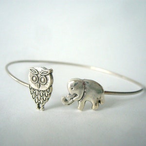 May include: A silver-toned bangle bracelet featuring an owl and an elephant charm. The owl charm has detailed feather and eye markings. The elephant charm has a raised trunk. The bracelet is a simple, elegant design.