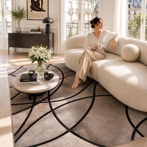 Puede incluir: Una alfombra beige con un diseño de líneas negras curvas. La alfombra está en un salón con un sofá blanco, una mesa de centro redonda y una obra de arte enmarcada en la pared. La habitación tiene grandes ventanas.