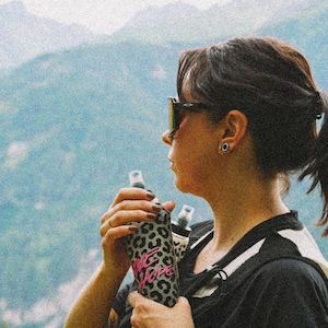 Peut inclure: Une housse pour bouteille d'eau à motif léopard gris et noir avec le texte rose "BIG HOPE" écrit en police cursive. La housse est conçue pour contenir une bouteille d'eau en plastique transparent avec un bouchon noir, offrant une prise en main.