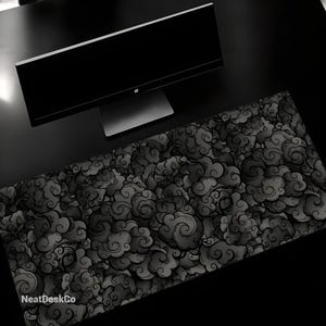 May include: Black and white desk mat with a repeating pattern of stylised clouds. The mat is on a black surface with a black computer monitor on top of it. The text "NeatDeskCo" is in the bottom left corner of the mat.