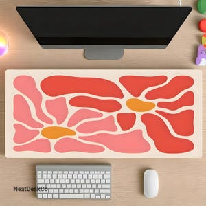 May include: A desk mat with a floral design featuring pink, red, and orange abstract shapes. The mat is cream-coloured and has a smooth surface. The text "NeatDeskCo" is printed in the bottom left corner.