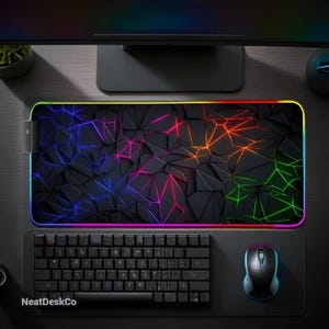May include: A black gaming mouse pad with a geometric design and a rainbow LED edge. A black keyboard and mouse are on the pad. The text "NeatDeskCo" is visible on the pad.