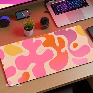 May include: A rectangular desk mat with a vibrant abstract design. The mat showcases a pattern of pink, orange, and yellow organic shapes on a beige background. The text "NeatDeskCo" is printed on the bottom left corner.
