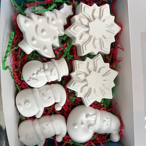May include: A collection of white, unpainted Christmas-themed plaster shapes, including a Christmas tree, snowflakes, and snowmen, nestled in red and green paper shred inside a blue box. Ideal for DIY holiday crafts.