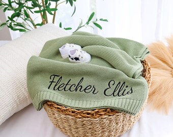 以下が含まれることがあります： 「Fletcher Ellis」と刺繍されたセージグリーンのニットベビーブランケットが、編みかごの中に置かれています。星のアクセントが付いた白いベビーシューズがブランケットの上にあります。白い枕と装飾用の花が背景にあります。