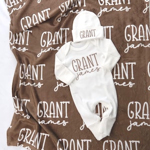 Op de afbeelding: Een baby cadeau set met een bruine deken met de naam "GRANT james" in witte letters. De set bevat een witte onesie en een bijpassende muts, beide met de naam "GRANT" in bruine letters. De deken is gemaakt van zacht materiaal.