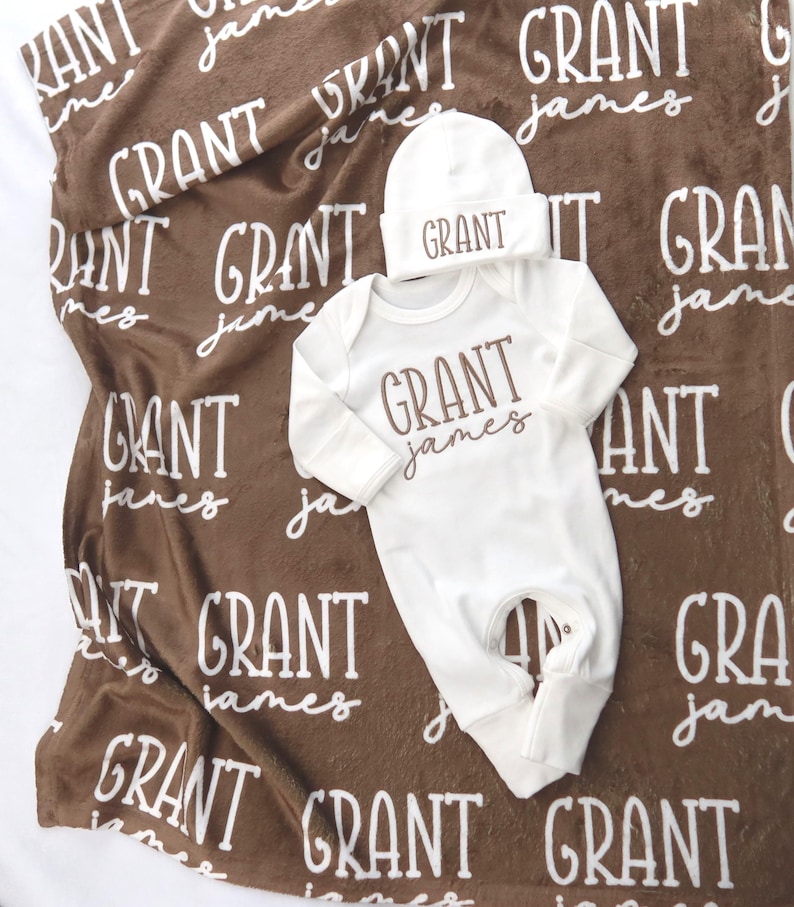 Op de afbeelding: Een baby cadeau set met een bruine deken met de naam "GRANT james" in witte letters. De set bevat een witte onesie en een bijpassende muts, beide met de naam "GRANT" in bruine letters. De deken is gemaakt van zacht materiaal.