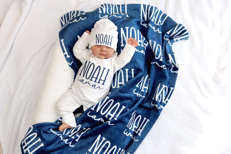 Op de afbeelding: Een pasgeboren baby in een witte onesie en muts, beide met de naam "NOAH" in marineblauw. De baby ligt op een marineblauwe deken met de naam "NOAH Lamar" in wit. De set ligt op een witte ondergrond.