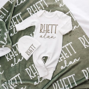 Op de afbeelding: Een baby cadeau set met een witte onesie, een bijpassende muts en een groene deken. De onesie en muts zijn bedrukt met de naam "RHETT alan" in bruin. De groene deken heeft de naam "RHETT alan" in het wit.