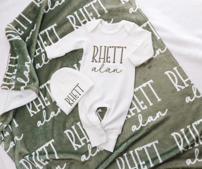 Op de afbeelding: Een baby cadeau set met een witte onesie, een bijpassende muts en een groene deken. De onesie en muts zijn bedrukt met de naam "RHETT alan" in bruin. De groene deken heeft de naam "RHETT alan" in het wit.