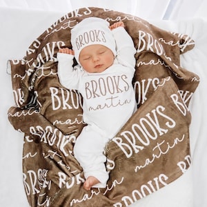 May include: A white long-sleeve infant bodysuit and matching beanie set, both personalized with the name BROOKS martin in brown text, resting on a brown plush blanket featuring a repeating white pattern of the name BROOKS martin throughout.