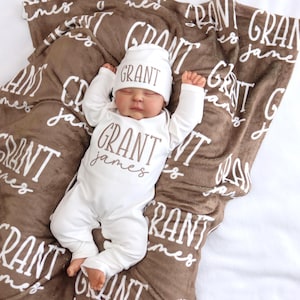 Op de afbeelding: Een pasgeboren baby in een witte onesie en muts, beide met "GRANT James" in bruin gedrukt. De baby ligt op een bruine deken met de naam "GRANT James" herhaald in wit. De deken ligt op een witte ondergrond.