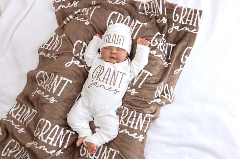 Op de afbeelding: Een pasgeboren baby in een witte onesie en muts, beide met "GRANT James" in bruin gedrukt. De baby ligt op een bruine deken met de naam "GRANT James" herhaald in wit. De deken ligt op een witte ondergrond.