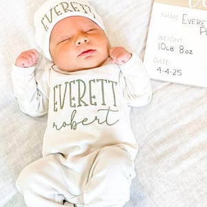 May include: A newborn baby in a white onesie and hat, both with the name "EVERETT" printed in green. The baby lies on a white sheet next to a wooden sign with the baby's name, weight (4.7 kg), and date of birth.
