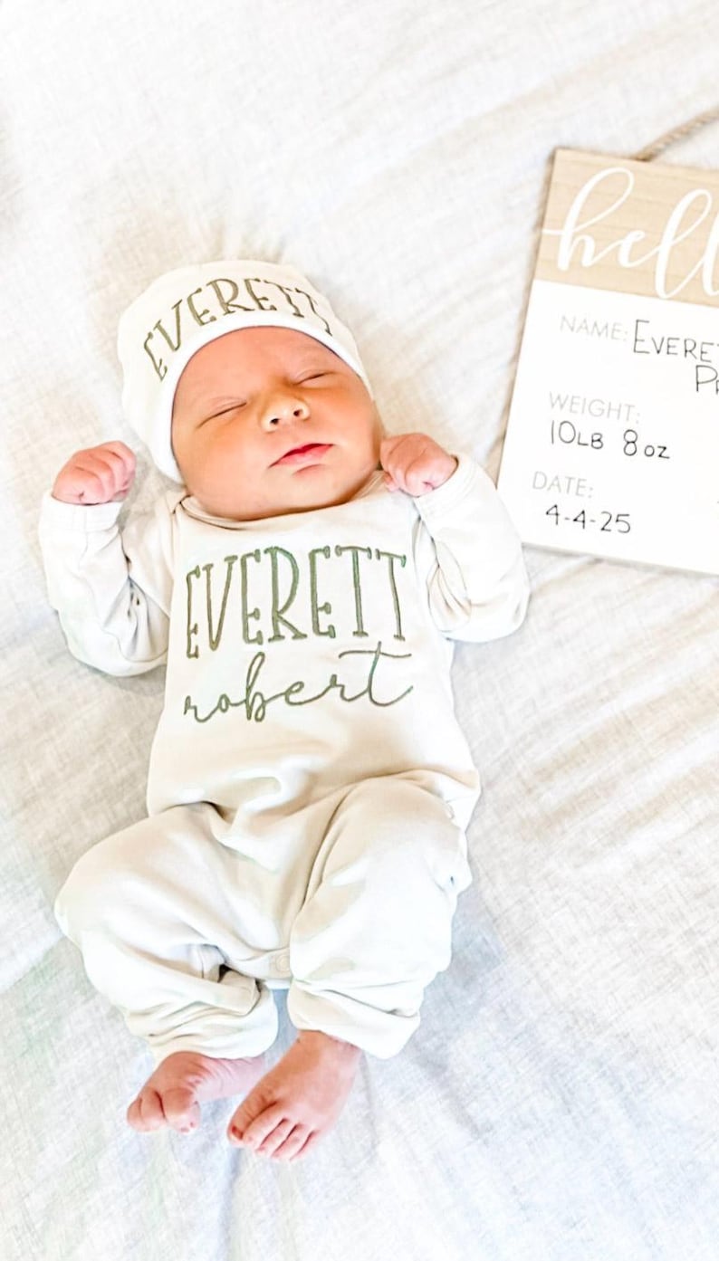 May include: A newborn baby in a white onesie and hat, both with the name "EVERETT" printed in green. The baby lies on a white sheet next to a wooden sign with the baby's name, weight (4.7 kg), and date of birth.