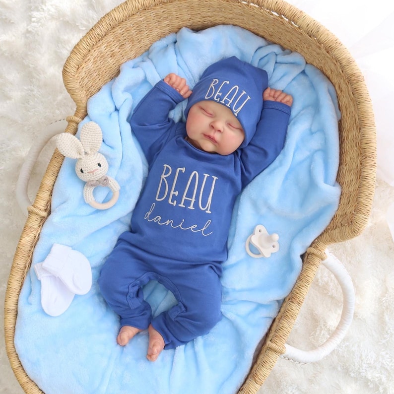 May include: A blue baby onesie with the name "Beau Daniel" embroidered on the front. The onesie is paired with a matching blue hat with the name "Beau" embroidered on it. The baby is sleeping in a wicker basket.