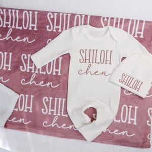 Op de afbeelding: Een baby cadeau set met een witte romper en muts met de naam "SHILOH" in een sierlijk lettertype. De set ligt op een oudroze deken met de naam "SHILOH" in een patroon.