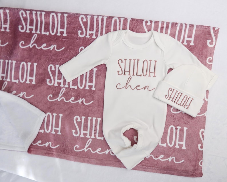 Op de afbeelding: Een baby cadeau set met een witte romper en muts met de naam "SHILOH" in een sierlijk lettertype. De set ligt op een oudroze deken met de naam "SHILOH" in een patroon.