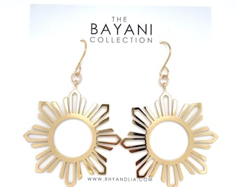 May include: A pair of gold-toned dangle earrings featuring a sunburst design with a central circular cutout. The earrings are attached to a white display card printed with the text THE BAYANI COLLECTION and WWW.RHYANDLIA.COM in black lettering.