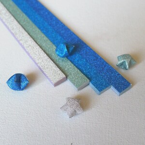 Puede incluir: Vista cenital de tiras de papel de origami y estrellas plegadas en tonos azules, plateados y verdes. Las tiras est&aacute;n dispuestas en diagonal, con varias estrellas de origami dispersas. El papel tiene un acabado brillante.