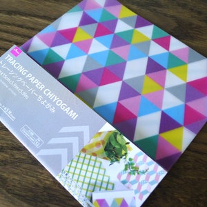 May include: A pack of tracing paper with a geometric pattern of pink, blue, yellow, green, and gray triangles. The pack is labeled "Tracing Paper Chiyogami" and includes 5 sheets of paper, each measuring 5.9 inches by 5.9 inches.