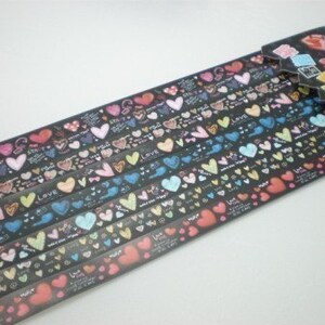 Puede incluir: Un rollo de cinta washi de colores con un patr&oacute;n repetitivo de corazones y la palabra "Love". La cinta est&aacute; sobre un fondo negro y tiene un borde blanco.