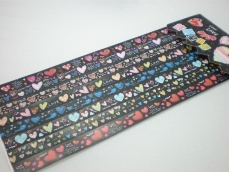 Puede incluir: Un rollo de cinta washi de colores con un patr&oacute;n repetitivo de corazones y la palabra "Love". La cinta est&aacute; sobre un fondo negro y tiene un borde blanco.